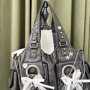 May include: Black leather shoulder bag with silver hardware, including buckles, rivets, and decorative charms. Features two front pockets, large silver rings, and white lace bows. The bag has a structured design with a top handle.