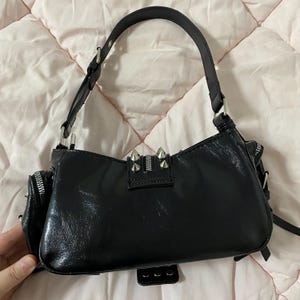 Puede incluir: Bolso de hombro negro con acabado brillante, cremalleras plateadas y detalles de tachuelas. Tiene un asa corta y un peque&ntilde;o bolsillo lateral. El bolso est&aacute; sobre una superficie acolchada rosa.