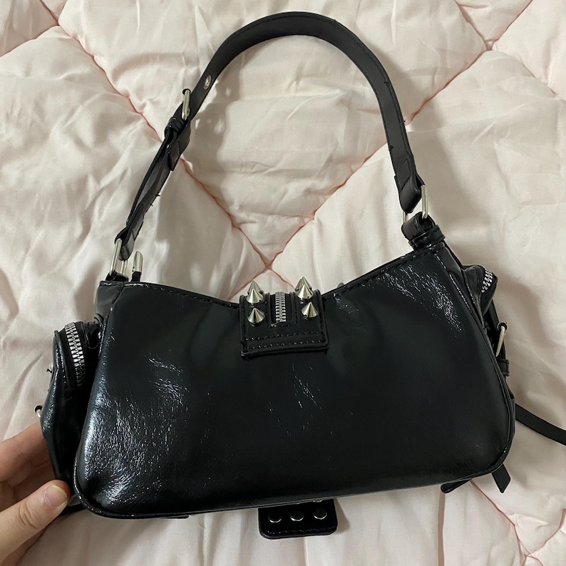 Puede incluir: Bolso de hombro negro con acabado brillante, cremalleras plateadas y detalles de tachuelas. Tiene un asa corta y un peque&ntilde;o bolsillo lateral. El bolso est&aacute; sobre una superficie acolchada rosa.