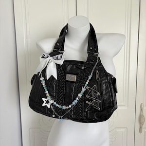 May include: Black leather handbag with a decorative white bow with zebra stripes, a beaded necklace, and safety pins. The bag has a braided design on the front and a silver metal plate.