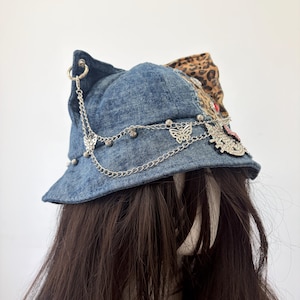 May include: A blue denim bucket hat featuring cat-ear points, a leopard print fabric panel, and silver-toned metal chains draped across the front. The hat is decorated with small silver butterfly charms, metal beads, and a rhinestone-encrusted cat face.