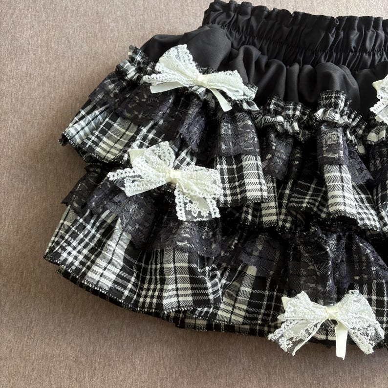May include: Black and white plaid skirt with multiple ruffled layers. The skirt features delicate white lace trim and is adorned with several white lace bows. The waistband is black and elasticized.
