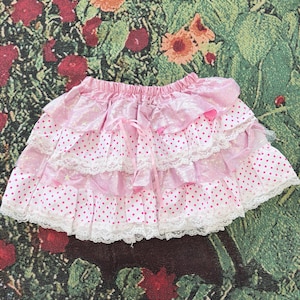 May include: A pink tiered skirt with white polka dots and lace trim. The skirt features a ruffled design, a satin ribbon bow, and star accents. The skirt is made of multiple layers of fabric.