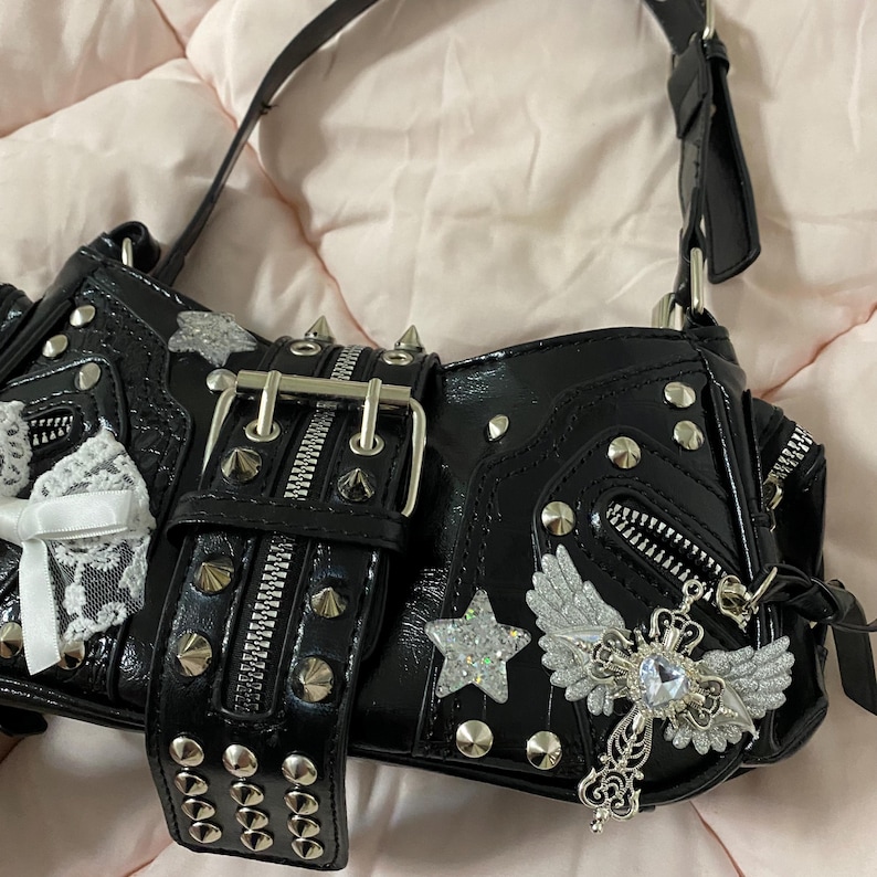 Puede incluir: Bolso de mano negro con herrajes plateados, tachuelas y cremalleras. Cuenta con una hebilla grande, una estrella y un colgante de cruz con alas. Incluye un lazo de encaje blanco en el lateral. El bolso tiene un acabado brillante.