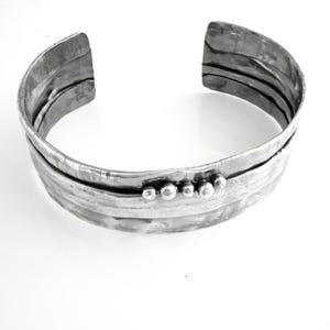 May include: A silver-toned cuff bracelet with a textured, layered design. The bracelet features a series of horizontal bands and small, round embellishments. The open-ended design allows for easy wear. This piece is a statement accessory.