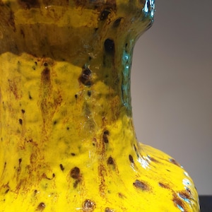May include: Close-up of a ceramic vase with a textured surface. The vase is primarily yellow with brown and dark brown accents. The glaze has a glossy finish, and the vase has a narrow neck and a wider body.
