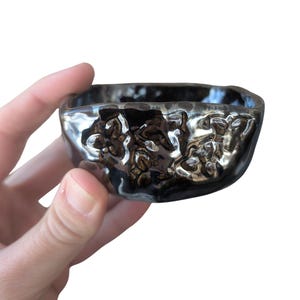 May include: A small, square, black ceramic bowl with a textured, metallic finish. The bowl has an uneven rim and a glossy, reflective surface. The interior appears smooth, contrasting with the exterior's rough, organic design.