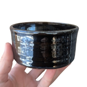 May include: A handmade, black ceramic bowl with a glossy finish. The bowl has a cylindrical shape with a slightly textured surface. The dark color and simple design suggest a rustic or minimalist aesthetic. The bowl is likely used for serving or display.