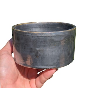 May include: A handmade, dark gray ceramic bowl with a cylindrical shape. The bowl has a slightly textured surface and a golden-brown rim. The bowl is held in a hand against a white background.