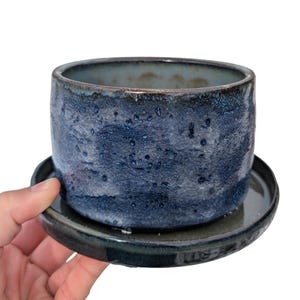 May include: A handcrafted ceramic planter with a deep blue glaze and speckled texture. The cylindrical pot sits on a matching saucer, creating a cohesive design. The planter is suitable for small plants and adds a touch of artistry to any space.