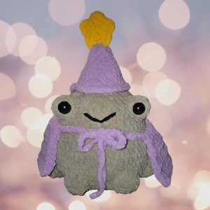 May include: A handmade plush frog with a purple hat and scarf, and a yellow star on top. The frog has large black eyes and a stitched smile. The plush toy is a soft, textured material.