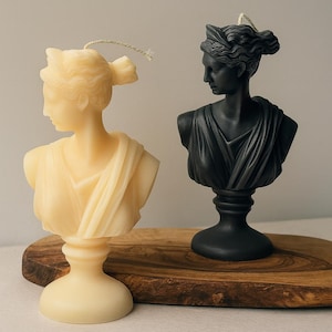 May include: Two bust candles, one cream-colored and one black, sit on a wooden board. The candles depict a classical female figure with detailed hair and draped clothing. Each candle has a wick.