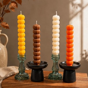 May include: Four tall, cylindrical pillar candles with a stacked, bubble-like texture in yellow, brown, cream, and orange. The candles are held in two clear green glass candle holders with a diamond pattern and two matte black pedestal candle holders.