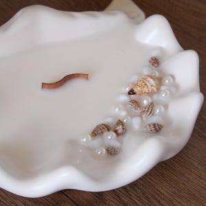 May include: White ceramic seashell shaped candle holder with a brown wooden wick and white pearls and seashells embedded in the wax.