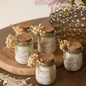 May include: Five small glass jars with cork lids filled with white candles. Each jar has a small, round, brown paper label with the text "Thank you!" and a small floral design. The jars are arranged on a wooden surface with dried flowers.