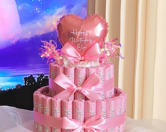 May include: A two-tiered gift tower constructed from pink Yogurette chocolate bars, wrapped with satin ribbons and bows. The top is decorated with a metallic pink heart-shaped balloon featuring the text Happy Birthday Ela in white script.