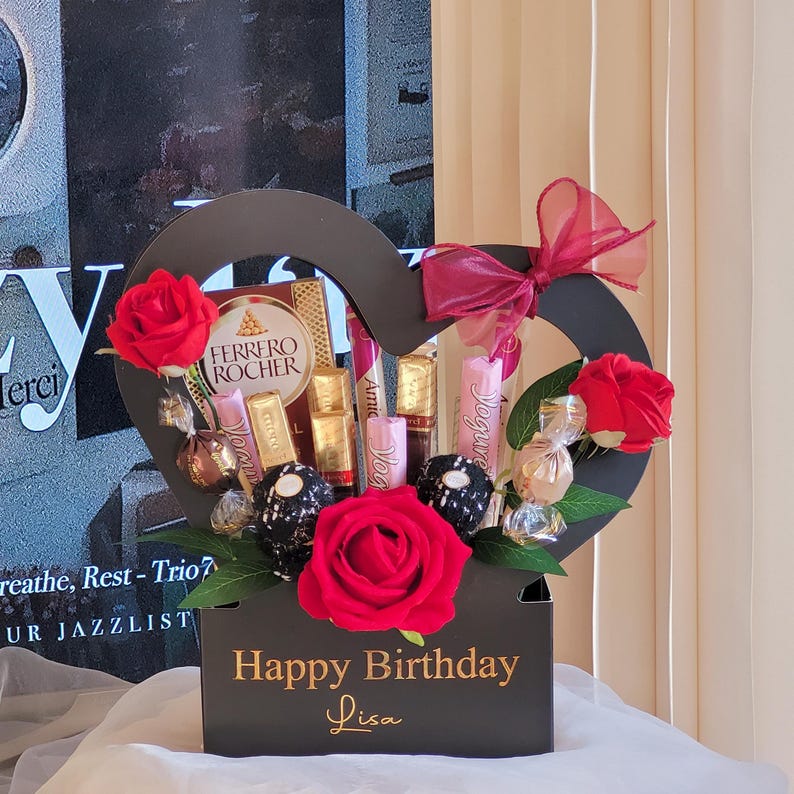 May include: A black heart-shaped gift box with a floral arrangement and chocolates. The box features red roses, a burgundy bow, and the text "Happy Birthday Lisa". The arrangement includes Ferrero Rocher chocolates and other wrapped candies.