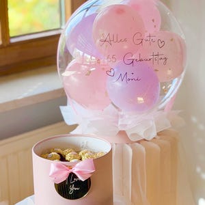 Könnte beinhalten: Ein transparenter Ballon gefüllt mit kleineren rosa und lila Ballons, beschriftet mit Alles Gute zum 65 Geburtstag Moni, verziert mit einer rosa Schleife. Daneben steht eine zylindrische rosa Geschenkbox mit gold verpackten Pralinen und einer Schleife.