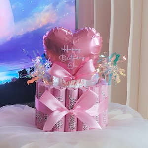 Könnte beinhalten: Ein zylindrisches Geschenkarrangement aus rosa Yogurette-Schokoriegeln, umwickelt mit einem rosa Satinband. Ein herzförmiger rosa Ballon mit der Aufschrift Happy Birthday Ela thront obenauf, ergänzt durch irisierendes Lametta und Raffaello-Kugeln.