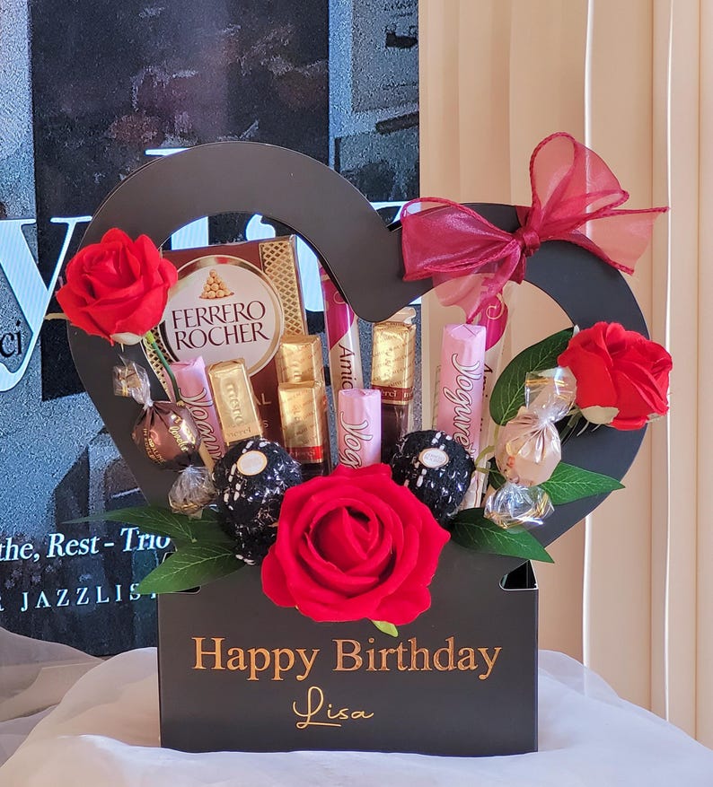 May include: A heart-shaped gift box filled with chocolates, candies, and red roses. The box is black with gold lettering that reads "Happy Birthday Lisa." A red bow adorns the top, creating a festive arrangement.