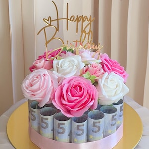 Könnte beinhalten: Eine kreisförmige Anordnung aus gerollten Fünf-Euro-Scheinen, die mit einem hellrosa Band befestigt sind, gekrönt von künstlichen weißen und rosa Rosen sowie einem goldenen Acryl-Tortentopper mit der Aufschrift Happy Birthday.