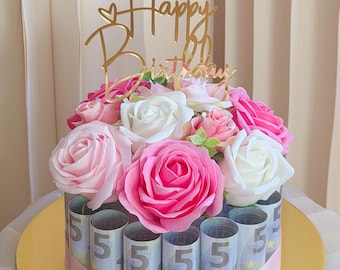 May include: A circular arrangement of rolled five-euro banknotes secured by a light pink ribbon, topped with a cluster of artificial white and pink roses and a gold-colored acrylic cake topper with the text Happy Birthday.