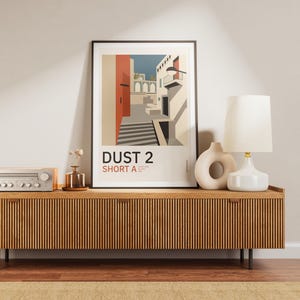 Può includere: Una stampa artistica verticale incorniciata intitolata DUST 2 SHORT A, che presenta un'illustrazione stilizzata di un vicolo urbano a tema desertico con scale, pareti rosse e cielo blu, esposta su una credenza in legno con una lampada.