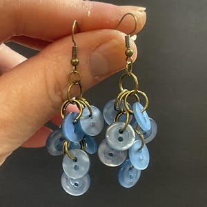 May include: A pair of dangle earrings with a cluster of light blue buttons hanging from gold-toned hoops. The earrings have a vintage style.