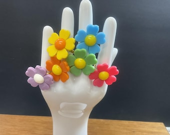 Op de afbeelding: Zes kleurrijke bloemvormige ringen op een witte handvormige display. De ringen zijn in tinten geel, oranje, groen, blauw, paars en roze.
