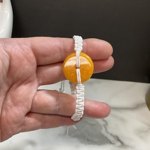 Peut inclure: Bracelet en macramé fait à la main avec une perle ronde orange et scintillante. La perle est maintenue par un cordon en macramé blanc, créant un accessoire unique et élégant. Le bracelet est conçu avec une esthétique simple mais élégante.