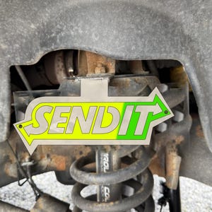 May include: A close-up of a vehicle's suspension system, featuring a coil spring, shock absorber, and various metal components. A sticker with the word "SEND IT" in a yellow and green arrow design is prominently displayed.