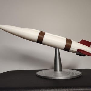 May include: A white and brown model rocket with a red tip and fins, displayed on a silver stand. The rocket has two brown bands around its body. The stand has a conical base and a circular platform.