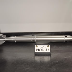 May include: A metallic silver B-61 MOD-12 bomb model with a pointed nose and finned tail. The model has a cylindrical body with engraved details and the text "W". A small sign displays "B-61 MOD-12".