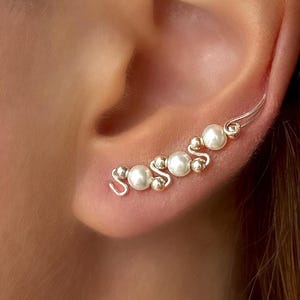 May include: A silver ear cuff featuring a series of faux pearls and small silver beads. The ear cuff curves along the ear, with the pearls and beads arranged in a decorative pattern. The jewelry is designed to be worn on the ear.