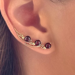 May include: Gold-toned ear climber earrings with three dark red gemstones. The earrings curve along the ear, with a delicate, leaf-like design. The gemstones are evenly spaced along the curve, creating a stylish and elegant look.