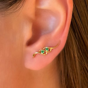 May include: A gold ear climber earring featuring a curved, flowing metal design set with two small, round, vibrant green gemstones. The piece follows the natural contour of the earlobe, showcasing a polished finish and a secure, sleek structure.