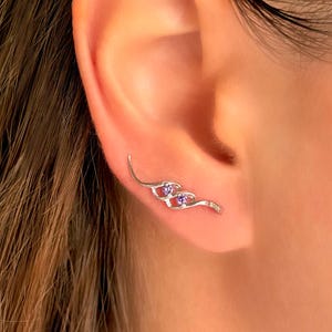 May include: A silver-toned ear climber earring featuring a curved, wave-like metal design set with two small, round, purple gemstones. The piece follows the natural contour of the earlobe, extending upward along the outer edge of the ear.