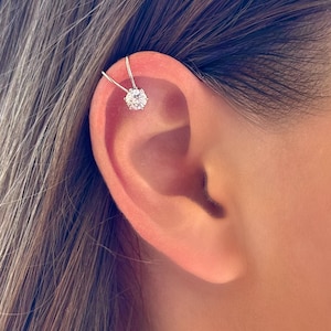 May include: A gold-colored ear cuff featuring a large, round, clear gemstone. The cuff curves around the ear, with a delicate bar extending to the top of the ear. The jewelry is set against a background of light brown hair and skin.