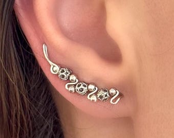 May include: A silver ear climber earring with a curved design. The earring features a series of small, textured beads and a delicate, curved shape that follows the ear's contour. The beads have a dark pattern.