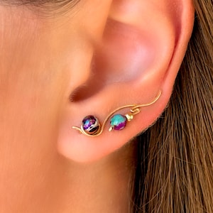 May include: Gold-toned ear cuff featuring two decorative, colorful beads. The first bead is a mix of purple, black, and white, while the second is teal and purple. The ear cuff has a delicate, curved design.