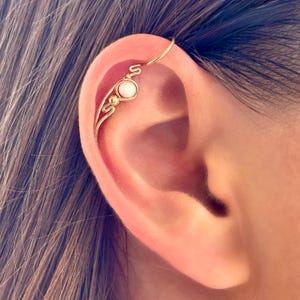 May include: A gold-colored ear cuff featuring a delicate design with a small, round, white gemstone. The cuff curves along the ear's outer edge, with decorative swirls and a small hoop at the top.