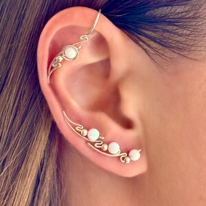 May include: Silver ear cuff with three white opal beads and a delicate, curved design. The ear cuff wraps around the ear, with a small opal bead at the top. The design is elegant and minimalist, suitable for everyday wear or special occasions.