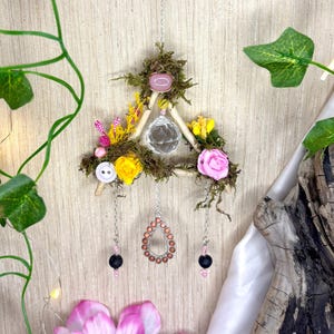 Puede incluir: Decoración de pared decorativa con un triángulo de madera adornado con musgo, flores y una bola de cristal transparente. El diseño incluye rosas rosas y amarillas, botones y colgantes con cuentas. También se ve un colgante plateado y un diseño floral.