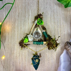 May include: A handmade wall hanging with a wooden triangle decorated with moss, green and white fabric roses, and a clear crystal ball. A gold chain suspends a green stone pendant and a beaded circle.