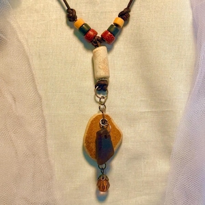 May include: A handmade necklace with a brown leather cord, adorned with yellow, green, and red beads. The pendant showcases a beige cylindrical bead, a tan stone, and a dark brown stone, completed with a small orange crystal bead.