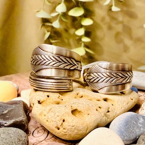 Könnte beinhalten: Drei silberne Ringe mit einem strukturierten Blattdesign sind auf einem hellbraunen Stein gestapelt. Die Ringe haben ein breites Band mit einem detaillierten Muster. Der Hintergrund zeigt eine verschwommene Ansicht von Grün und eine neutral getönte Oberfläche.