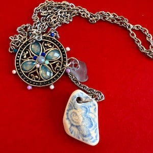 May include: Silver-toned necklace with a large, ornate pendant featuring a floral design with light blue and clear gemstones. The necklace includes a chain and a ceramic pendant with a blue and white pattern. The background is red.