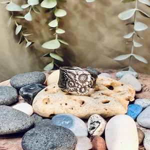 May include: A silver ring with a floral design. The ring is crafted from a spoon and features detailed flower and leaf patterns. The ring is displayed on a light-coloured rock surrounded by various stones.