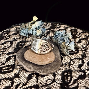 May include: A silver-toned metal ring crafted from a repurposed spoon handle, featuring an embossed decorative scroll pattern on the outer surface. The ring is positioned on a smooth, flat, oval-shaped stone, showcasing its curved, band-like structure.