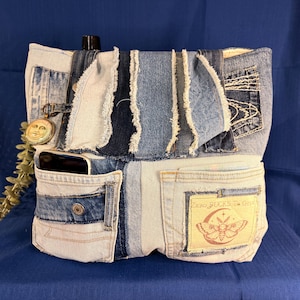 May include: A tote bag constructed from various shades of blue denim fabric patches with frayed edges. The bag features multiple exterior pockets, a circular moon face charm, and a rectangular patch with a moth graphic and the text ZERO FUCKS TO GIVE.